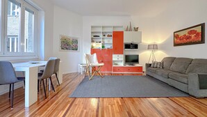 Una sala de estar moderna con piso de madera, un sofá gris, un escritorio blanco con sillas, un mueble de entretenimiento rojo y un cuadro en la pared.