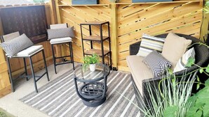 Un patio al aire libre con techo de madera, un sofá de mimbre, dos taburetes de bar, una mesa redonda de cristal y plantas en macetas.