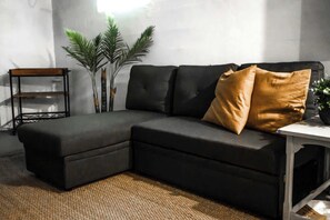 A sectional sofa with a chaise, a small table, and a plant in a modern room with a concrete wall and a woven rug.