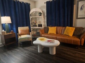 A living room with a brown leather sofa, a white coffee table with a flower, a wicker chair, a small side table with a lamp, and a white shelving unit.