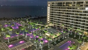 A hotel with multiple floors, a pool area with lounge chairs, and a view of the ocean.