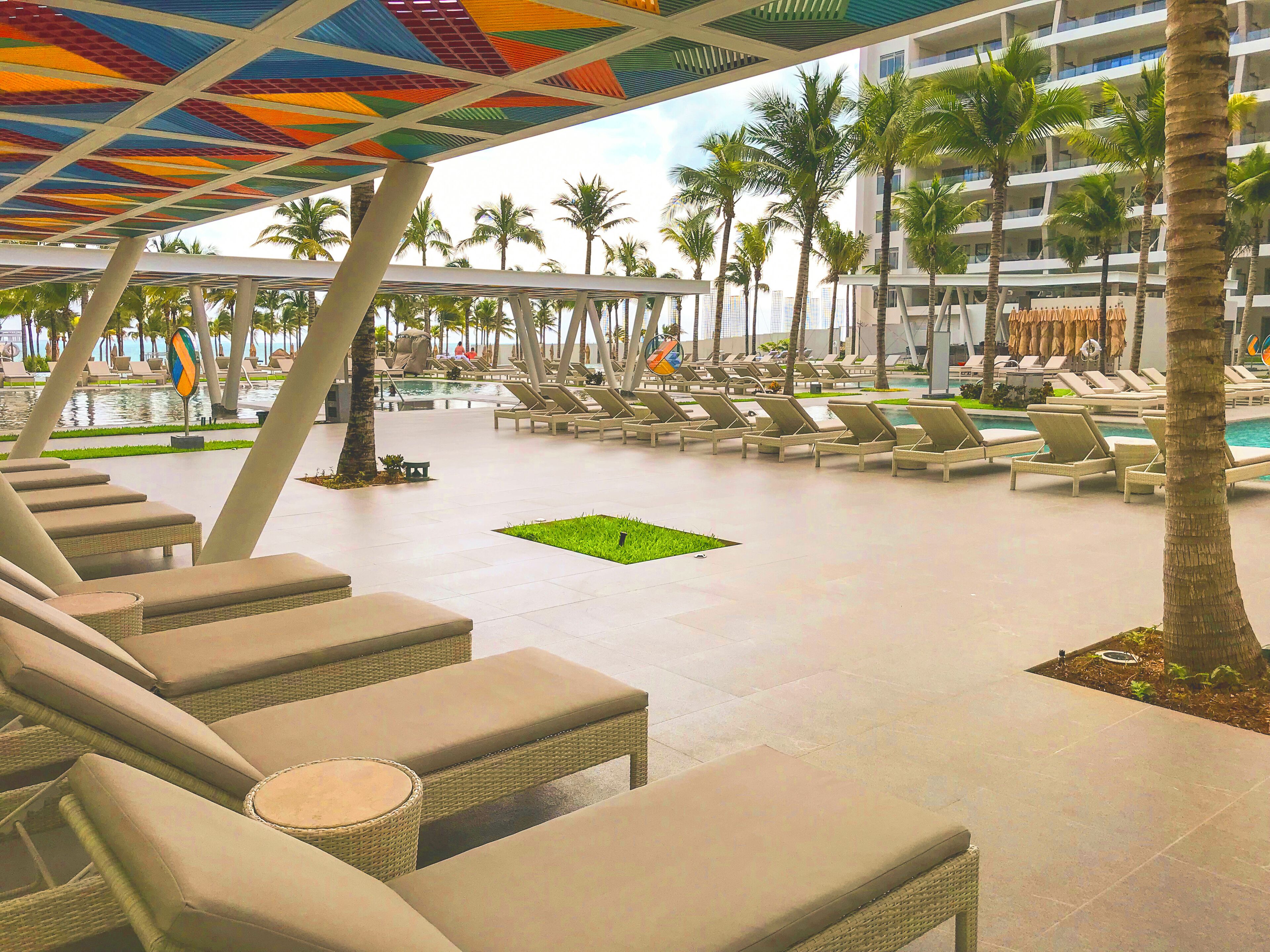 A resort pool area with lounge chairs, palm trees, and a colorful ceiling structure.