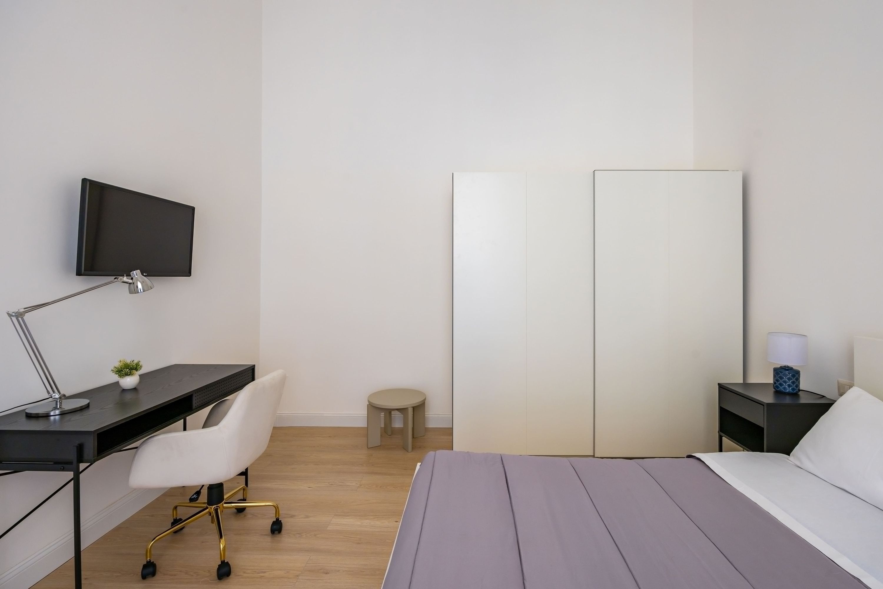 A bedroom with a bed, a desk with a chair, a TV, a nightstand, and a small stool.