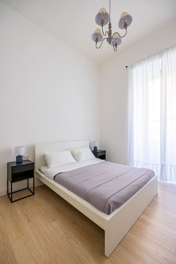 A minimalist bedroom with a white bed, two bedside tables, a chandelier, and sheer curtains.