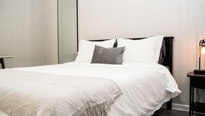 A neatly made bed with white linens and a grey pillow, a bedside table with a lamp, and a wall with vertical lines.
