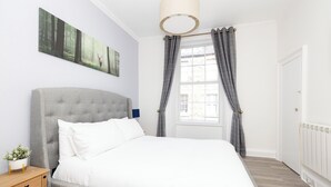 A bedroom with a grey upholstered bed, white bedding, a bedside table with a plant, a large window with grey curtains, and a wall-mounted artwork of a forest scene. - Argyl | Charming 2-Bedroom Apt. on Thistle Street (Edinburgh)