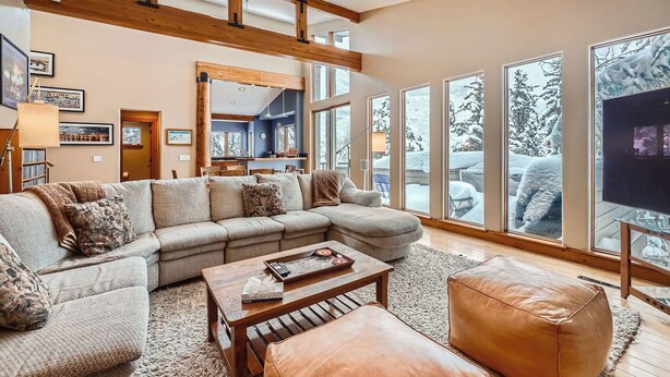 Una sala de estar acogedora con un sofá modular, una mesa de centro y un reposapiés de cuero. Amplias ventanas ofrecen vistas a árboles cubiertos de nieve.
