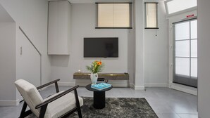 Una sala de estar moderna con un sofá, una mesa de centro con un florero y un libro, un televisor de pantalla plana montado en la pared y un ventanal con persianas.