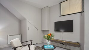 Una sala moderna con un sofá blanco, una mesita redonda con un libro y flores, un televisor montado en la pared y una escalera.