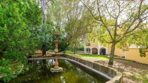 A serene garden with a fountain, lush greenery, and a building with arched windows.