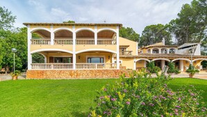 A two-story building with a stone base and yellow walls, featuring balconies and a garden with purple flowers.