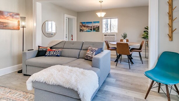 Un salón con un sofá gris, un otoman blanco y mullido, y una silla azul. Hay una zona de comedor con una mesa y sillas, y una cocina al fondo.