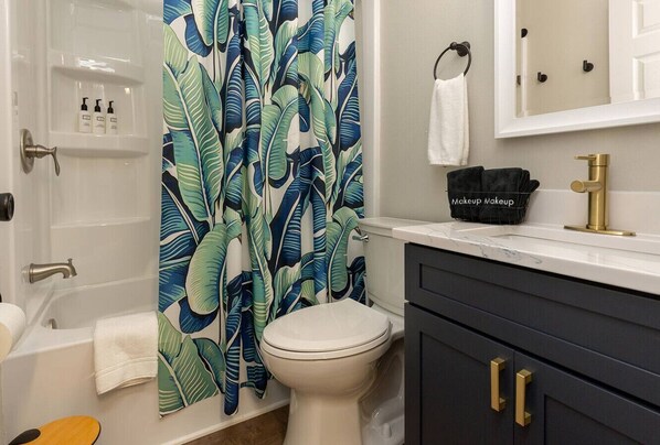 A bathroom with a toilet, a bathtub, a shower curtain with a tropical leaf pattern, a vanity with a black sink, and a gold faucet.