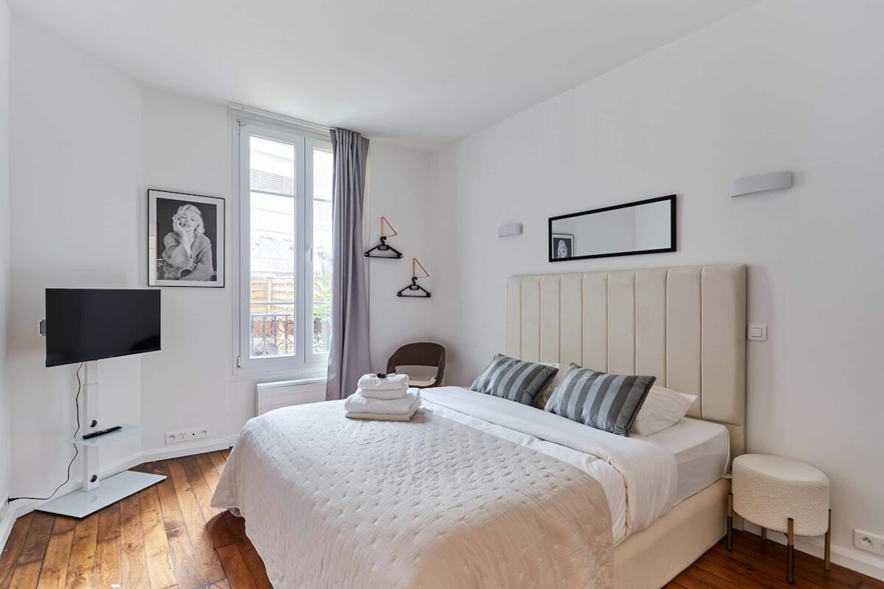 A bedroom with a bed, a TV, a window with curtains, a framed picture, and a bedside table.