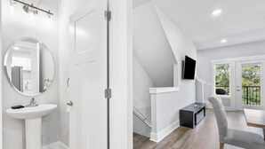 Un baño moderno con lavamanos y espejo blancos, una puerta y una escalera que lleva a un balcón con un televisor.