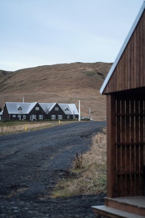 A row of dark wooden buildings with a gravel road in front, set against a hilly backdrop. - Paradise Cottages (Vik I Myrdal)