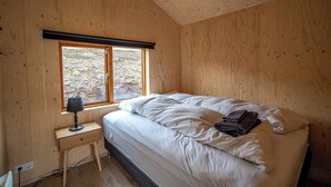 A bedroom with a wooden ceiling, a bed with white bedding, a small bedside table with a lamp, and a window with a view of the outdoors. - Paradise Cottages (Vik I Myrdal)