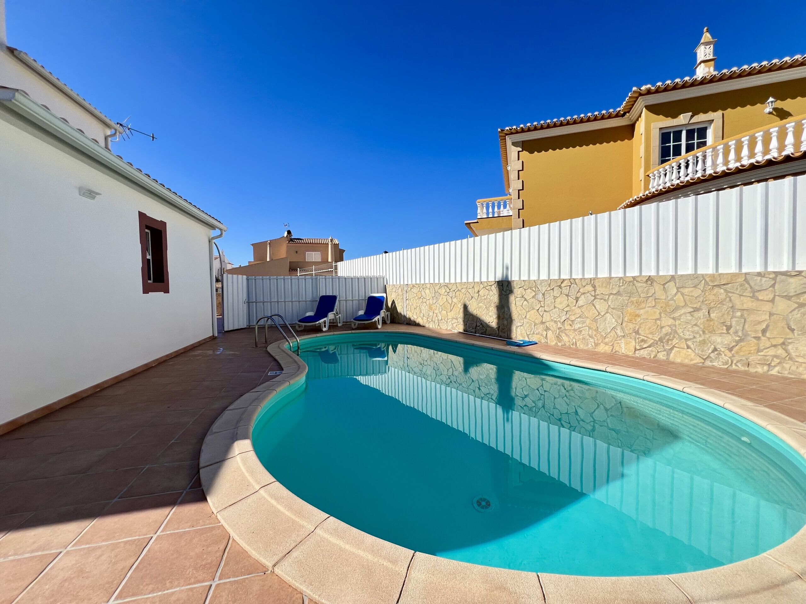 A swimming pool with two lounge chairs, a stone wall, and a white fence.