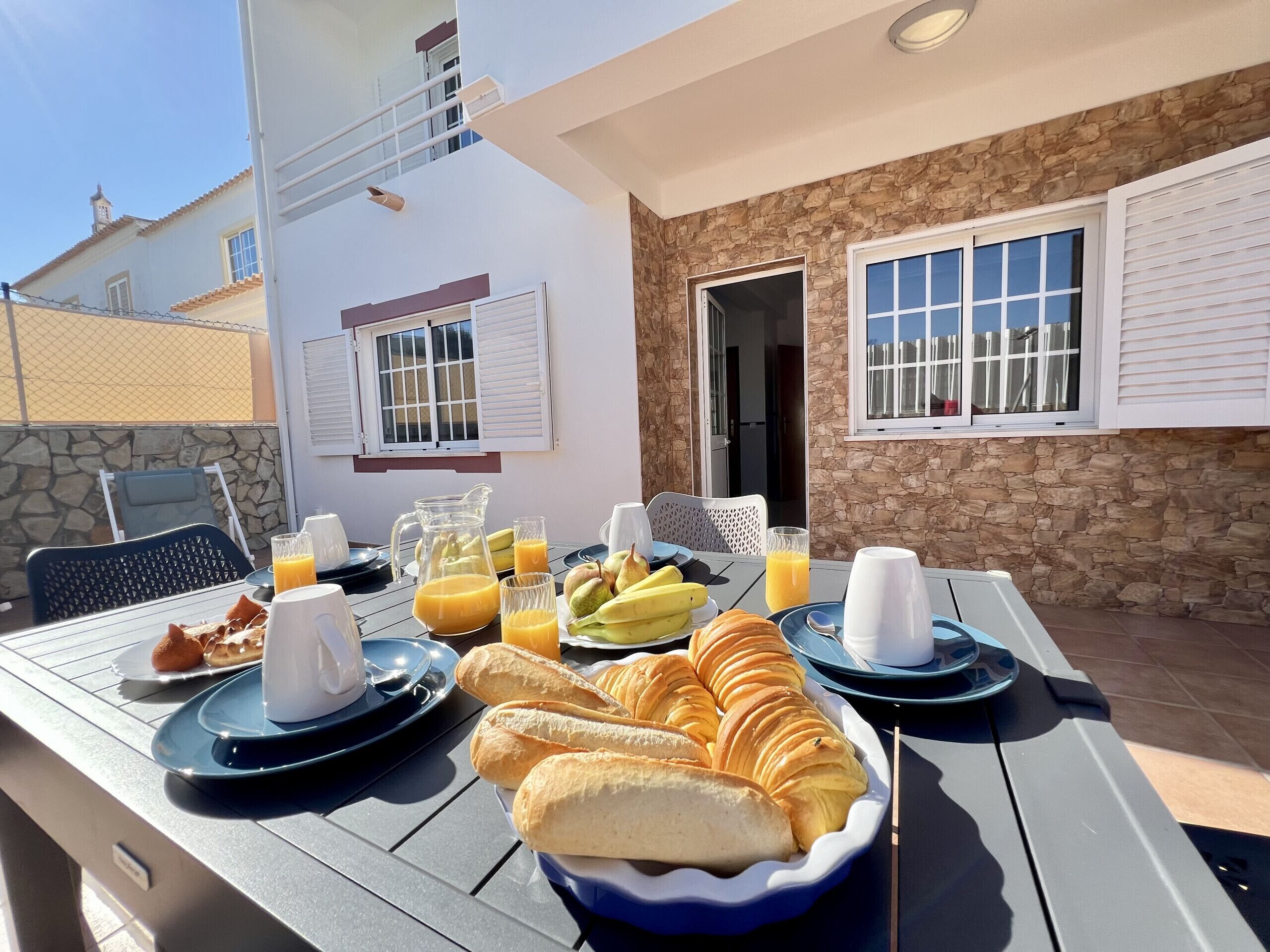 An outdoor dining area with a table set for breakfast, including croissants, bananas, and orange juice.