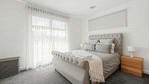 A modern bedroom with a tufted headboard, a large bed with multiple pillows, a bedside table with a lamp, and sheer curtains on the window. - Inlet Paradise in Rhyll (Rhyll)