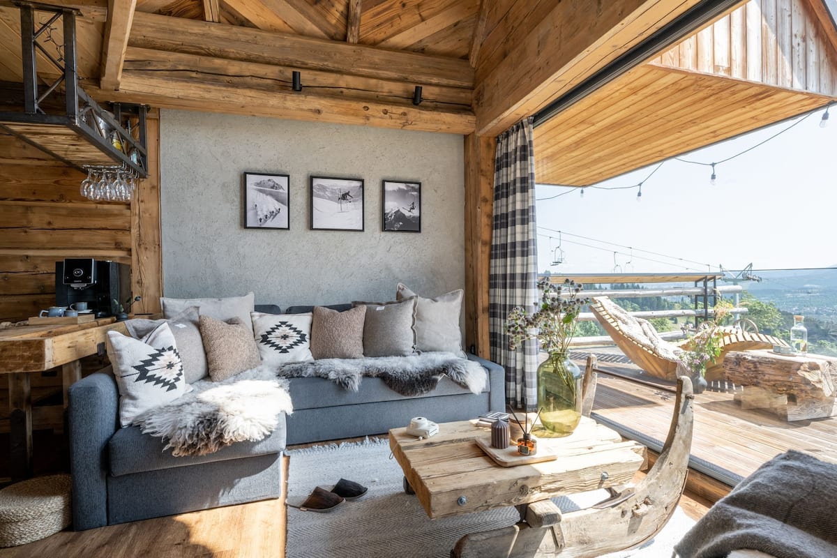 Ein gemütliches Blockhauss-Interieur mit Holzbalkendecke, einem grauen Sofa mit gemusterten Kissen, einer Kaffeemaschine und Blick ins Freie durch große Fenster.