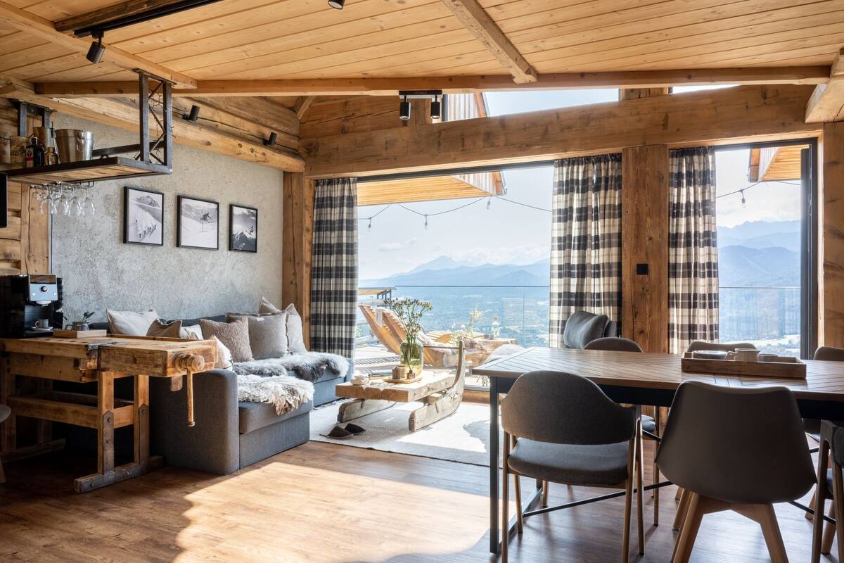 Ein gemütliches Blockhauss-Interieur mit Holzbalkendecke, einer Couch, einem Esstisch und Bergblick durch große Fenster.