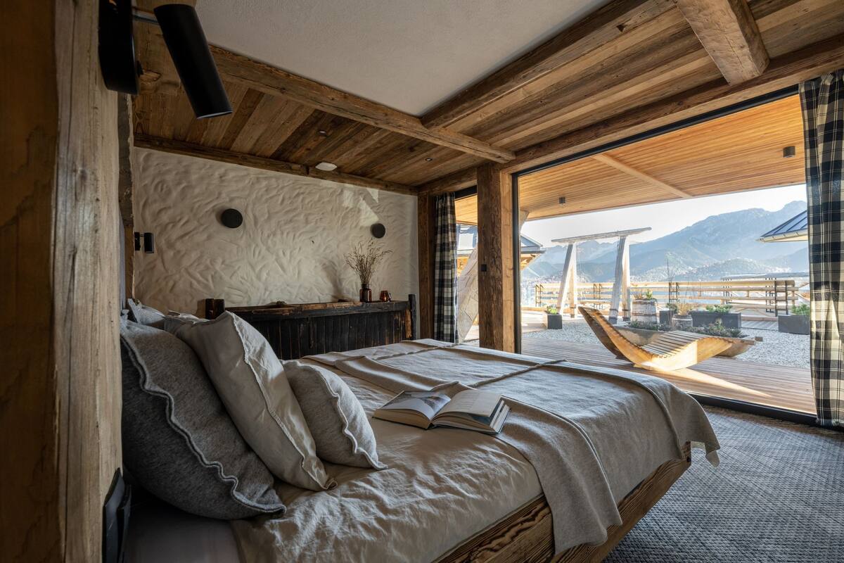 Ein Schlafzimmer mit einem großen Bett, Holzbalkendecke und Blick auf Berge durch eine Glastür.