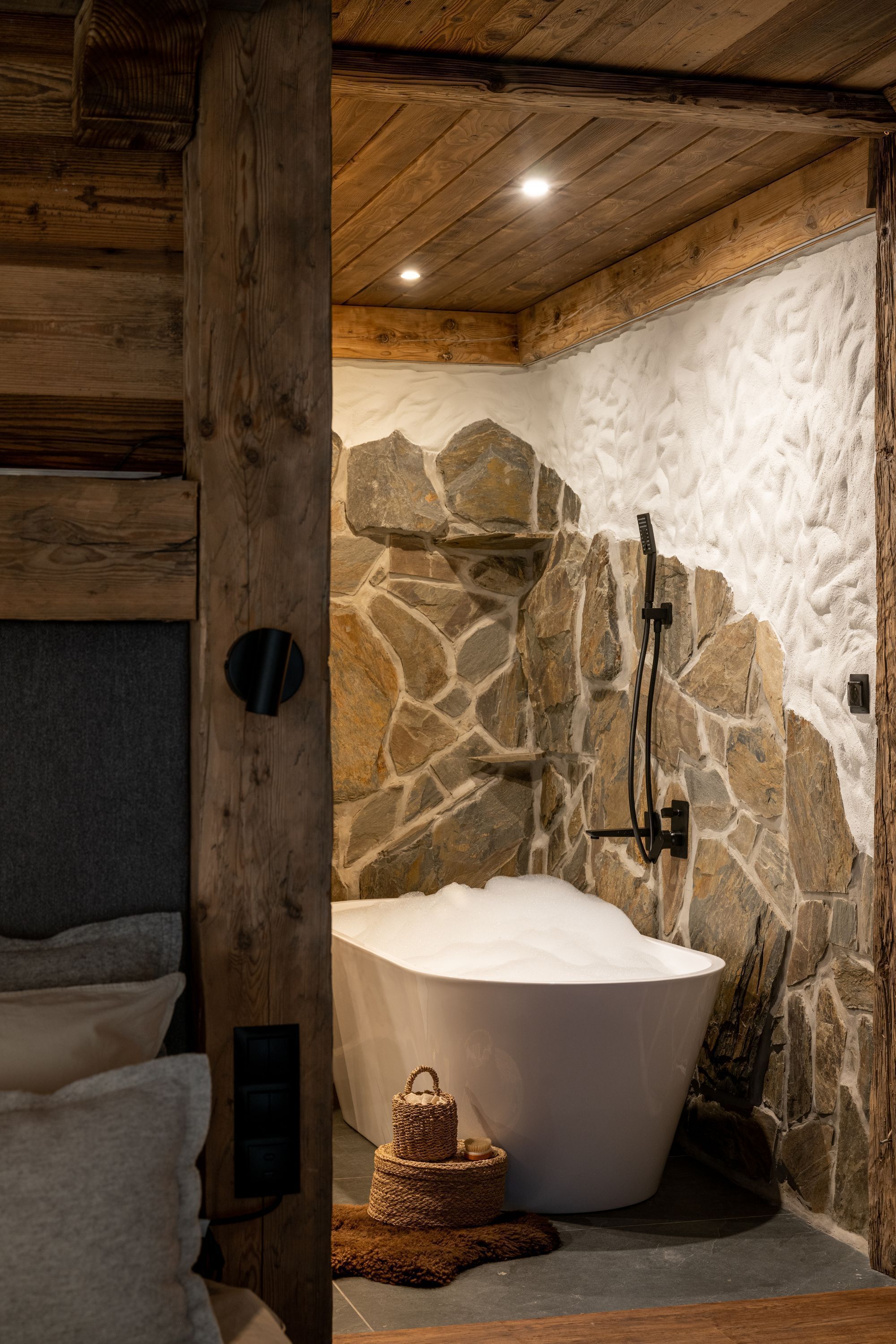 Un baño con paredes de piedra, una bañera independiente, vigas de madera y un muro de piedra.