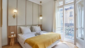 A hotel room with a large bed, a view of the city through the window, and a small side table with a lamp. - 4 Rooms Apartment Opera Vendome (Paris)