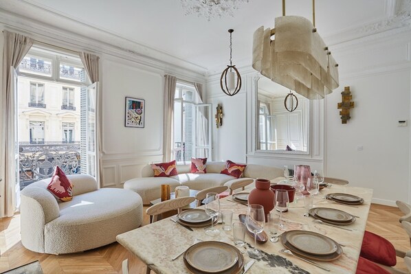 Un espace repas avec une longue table dressée pour un repas, entouré de canapés et de fauteuils, et offrant une vue sur les bâtiments à l’extérieur.