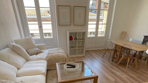 En moderne stue med sofa, sofabord i glas, spiseplads med bord og stole, samt store vinduer med udsigt til bygninger.