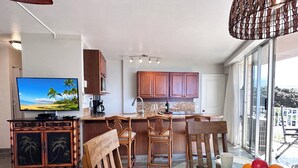 A modern kitchen with wooden cabinets, a dining area with a glass table, and a large window offering a view of the outdoors. - Nestled Between Two Beaches Kihei Oceanview 2BR Condo Panoramic Views Pool (Kihei)