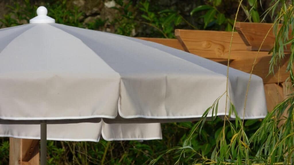 A white outdoor umbrella with a rounded top, set against a backdrop of greenery and wooden structures.