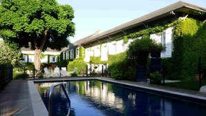 A swimming pool with a wooden deck, surrounded by lush greenery and white buildings with shutters. - La Tonnellerie (Tavers)