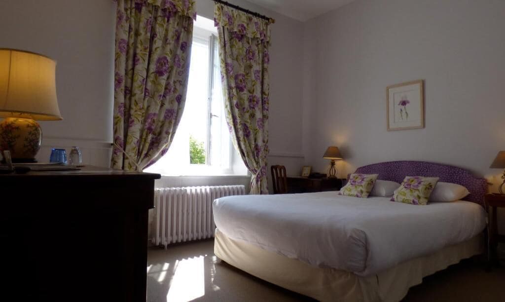 A bedroom with a bed, two bedside tables, a lamp, a radiator, and a window with curtains.