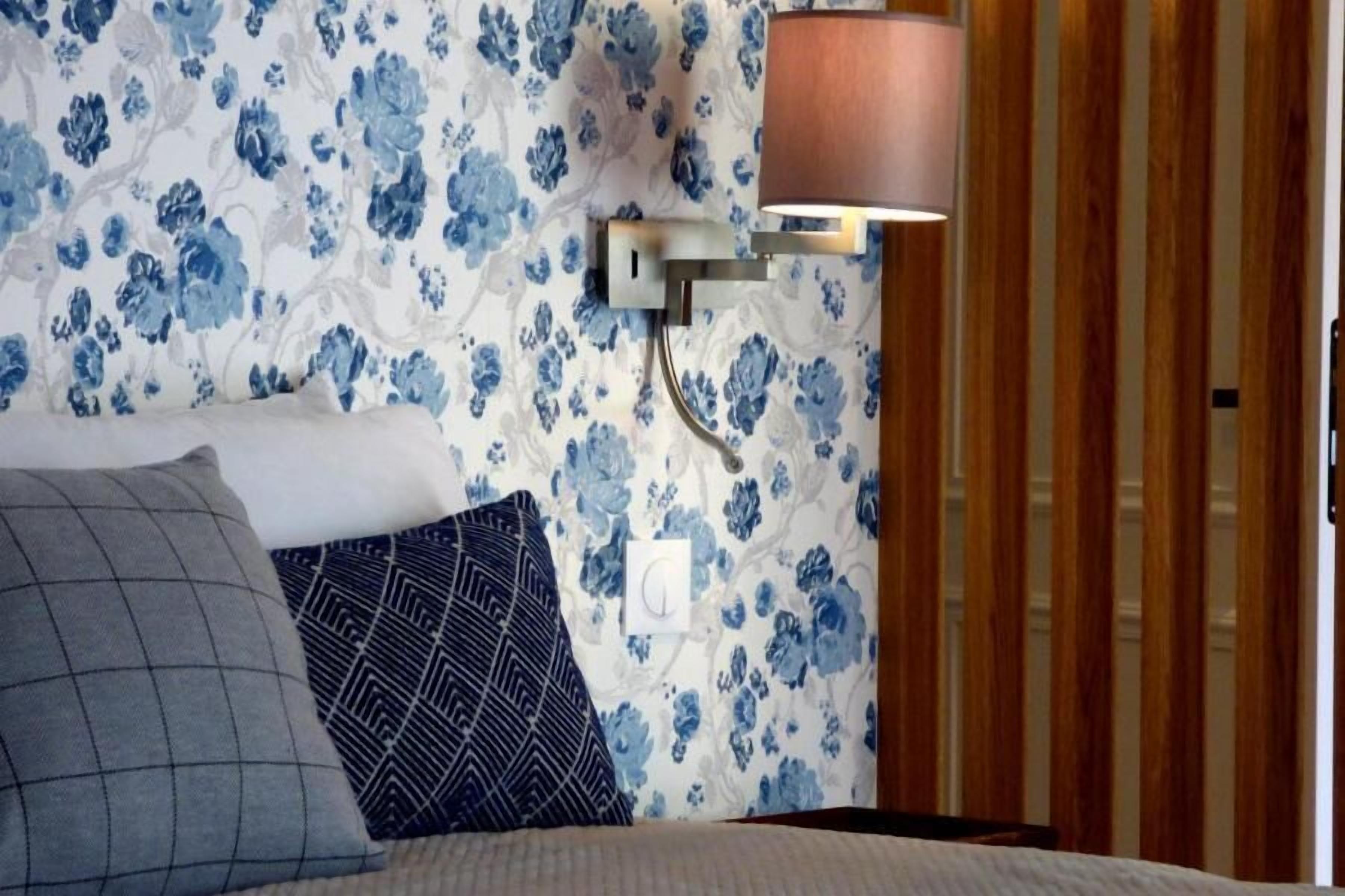 A bedroom with a floral wallpaper, a bed with two pillows, a wall-mounted lamp, and wooden paneling.