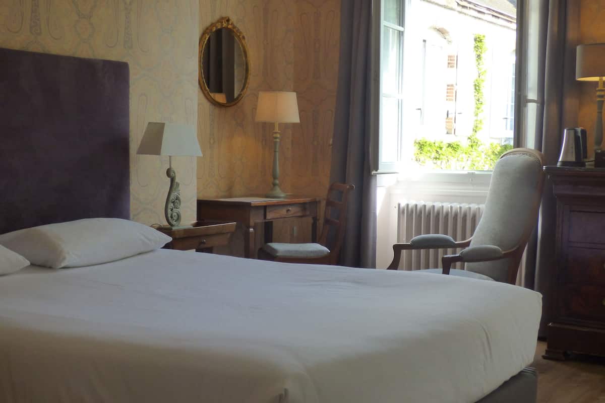 A hotel room with a large bed, a desk with a lamp, a chair, and a window with a view of buildings.