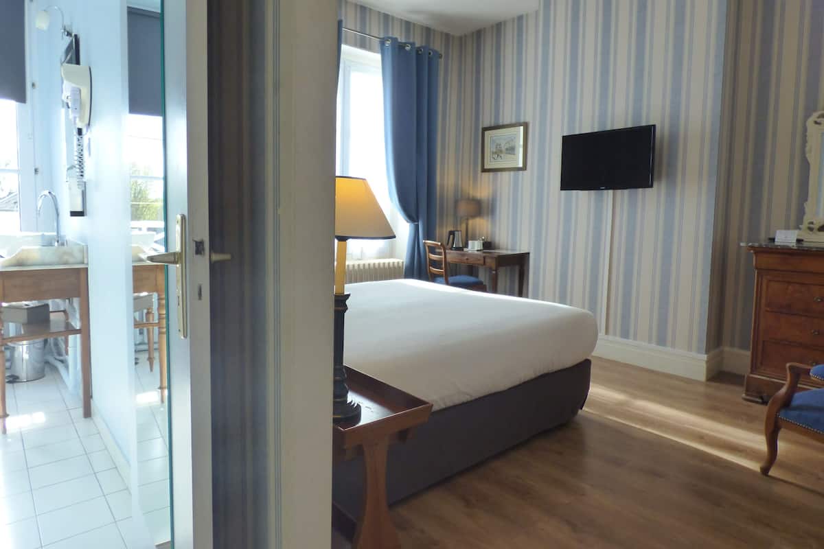 A hotel room with a bed, a desk, a chair, a TV, and a bathroom visible through an open door.