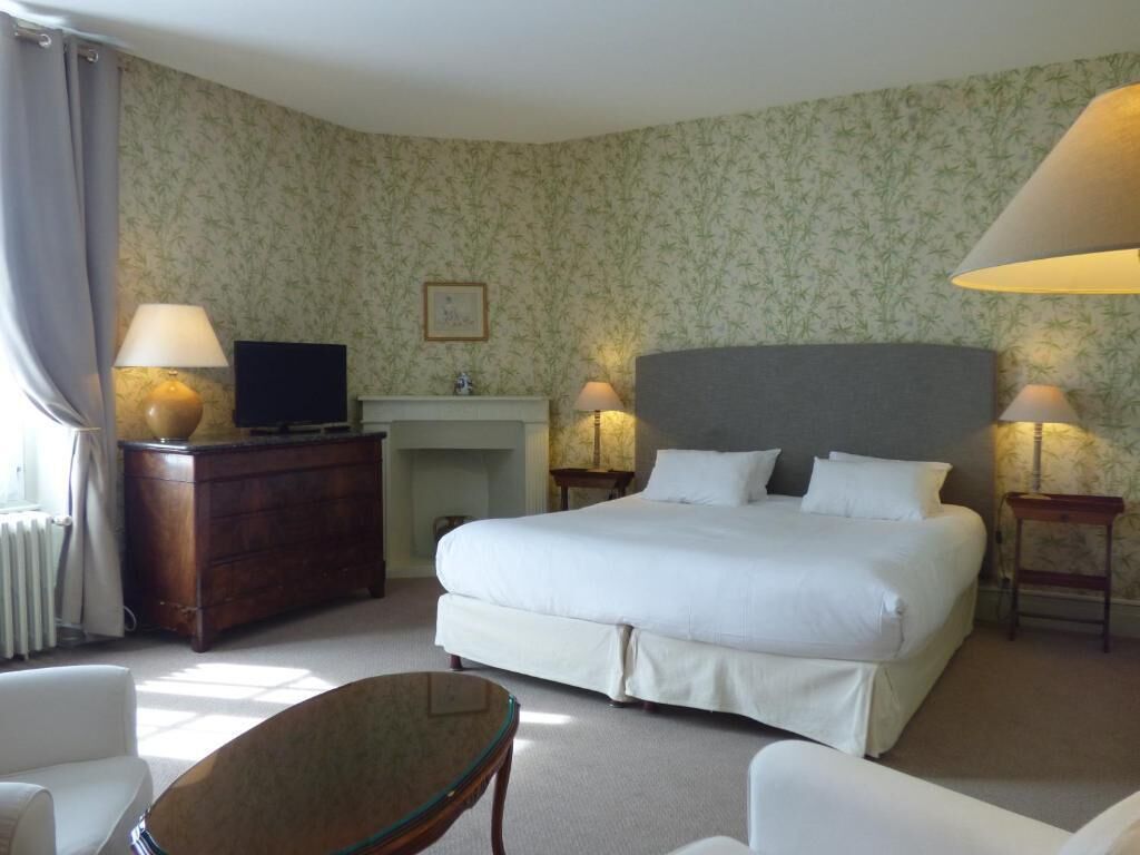 A hotel room with a large bed, a desk with a TV, two bedside lamps, a fireplace, and a small table.