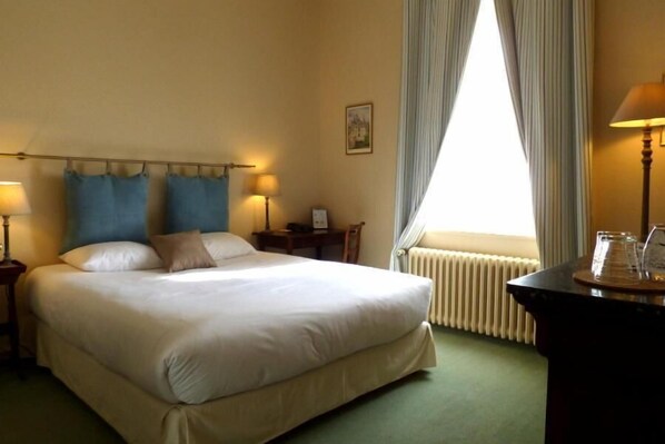 A hotel room with a large bed, two bedside lamps, a small table with a lamp, a window with curtains, and a radiator.