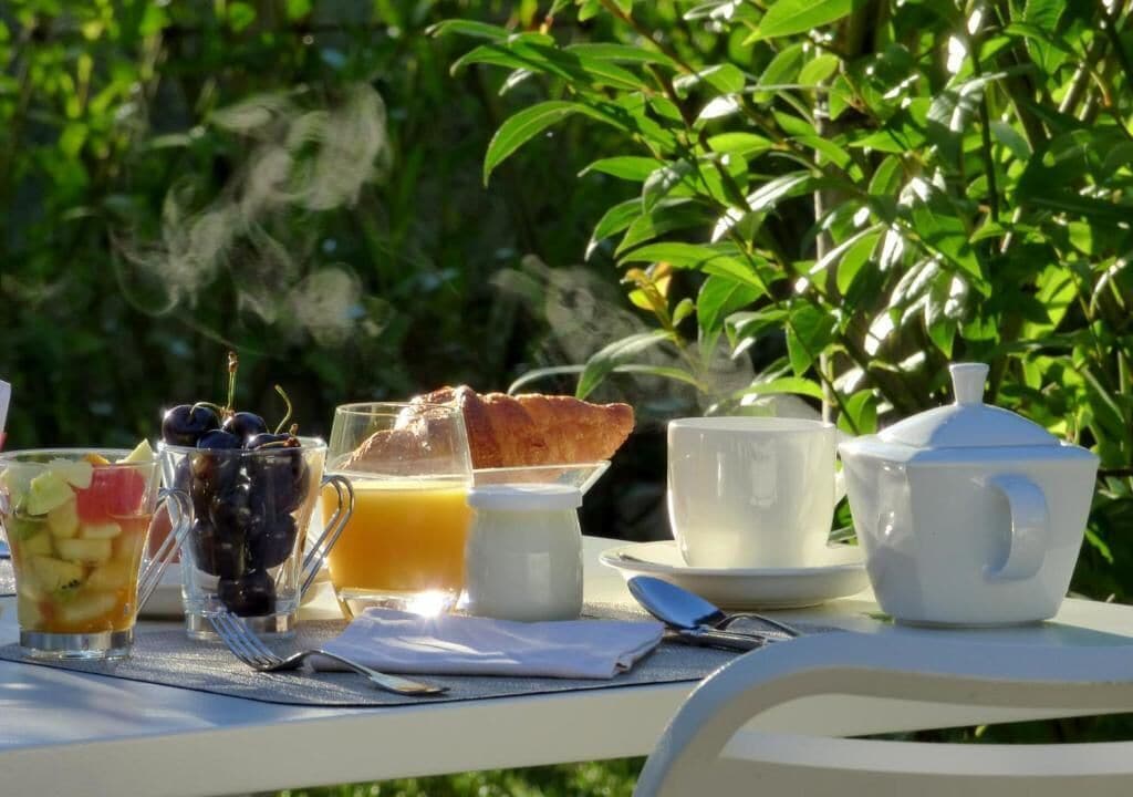 A breakfast table with fruit, juice, and pastries, set outdoors with greenery in the background.