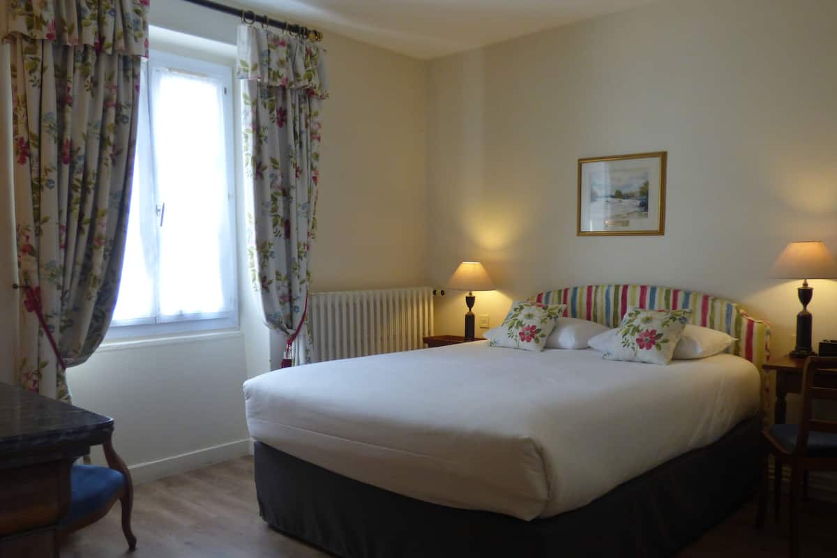 A neatly arranged bedroom with a large bed, two bedside lamps, a radiator, and a window with floral curtains.