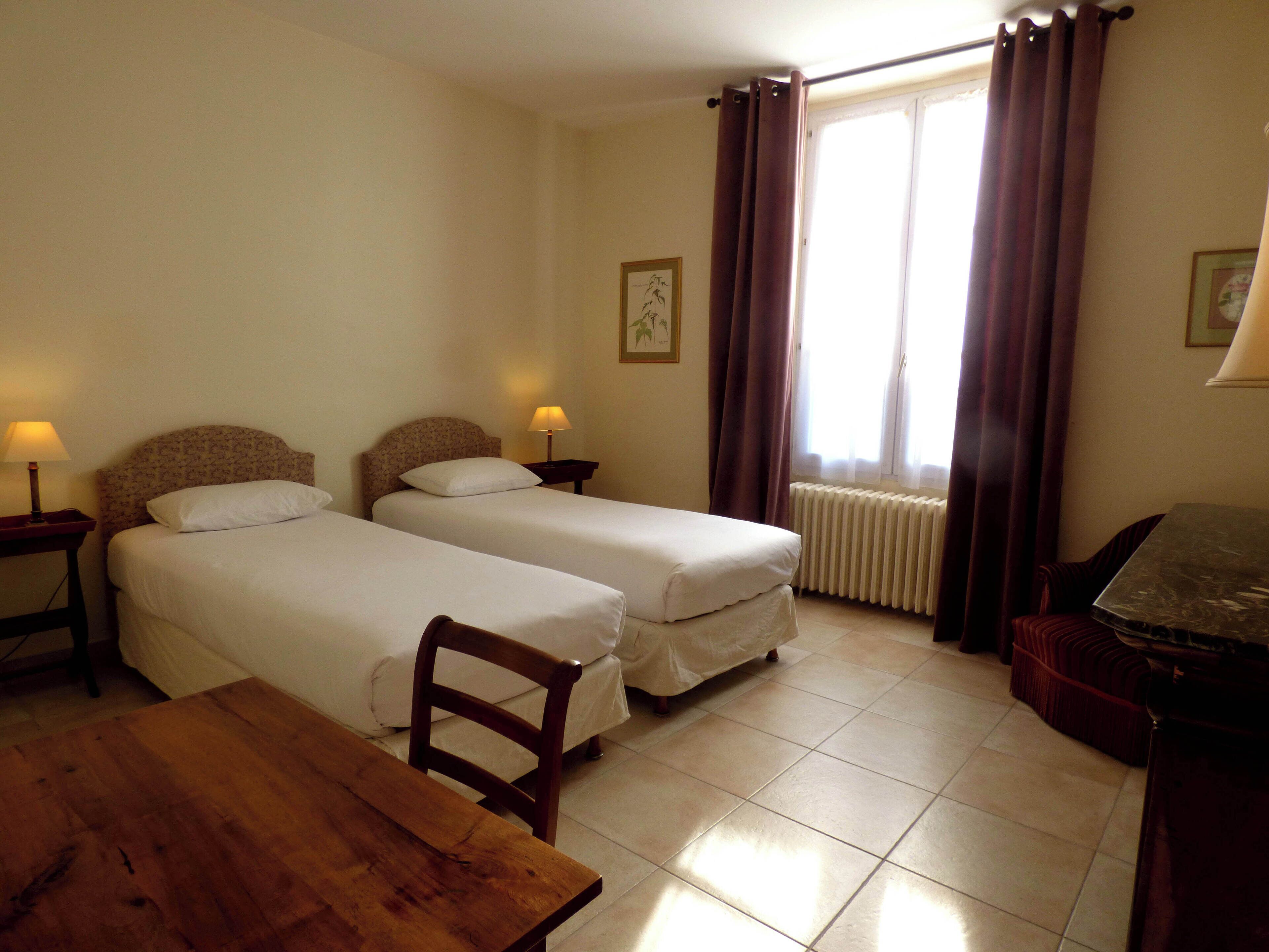 A hotel room with two single beds, a wooden table, a chair, a desk, and a window with curtains.