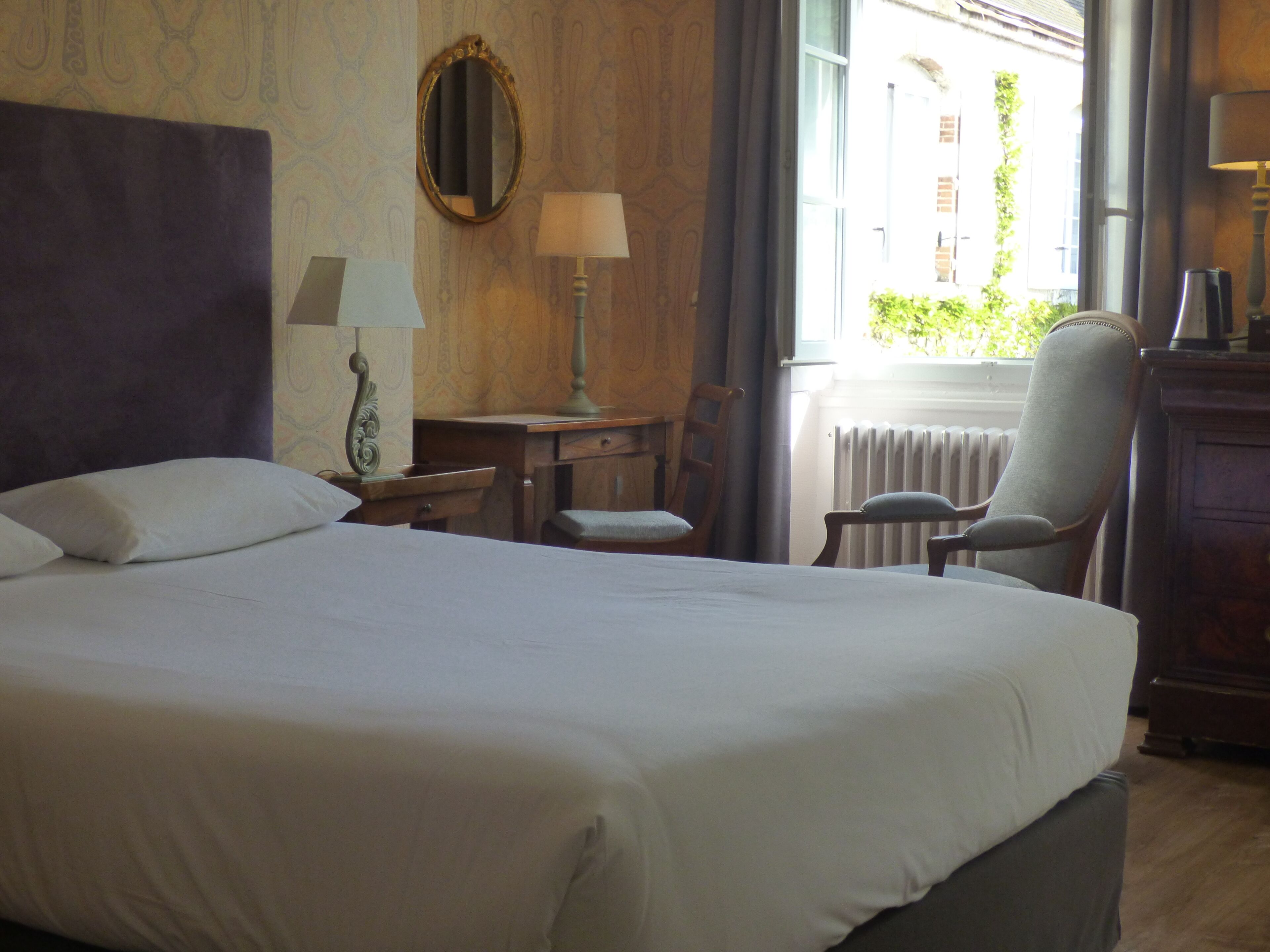 Quarto de hotel com uma cama grande, uma escrivaninha com luminária, uma cadeira e uma janela com vista para edifícios.