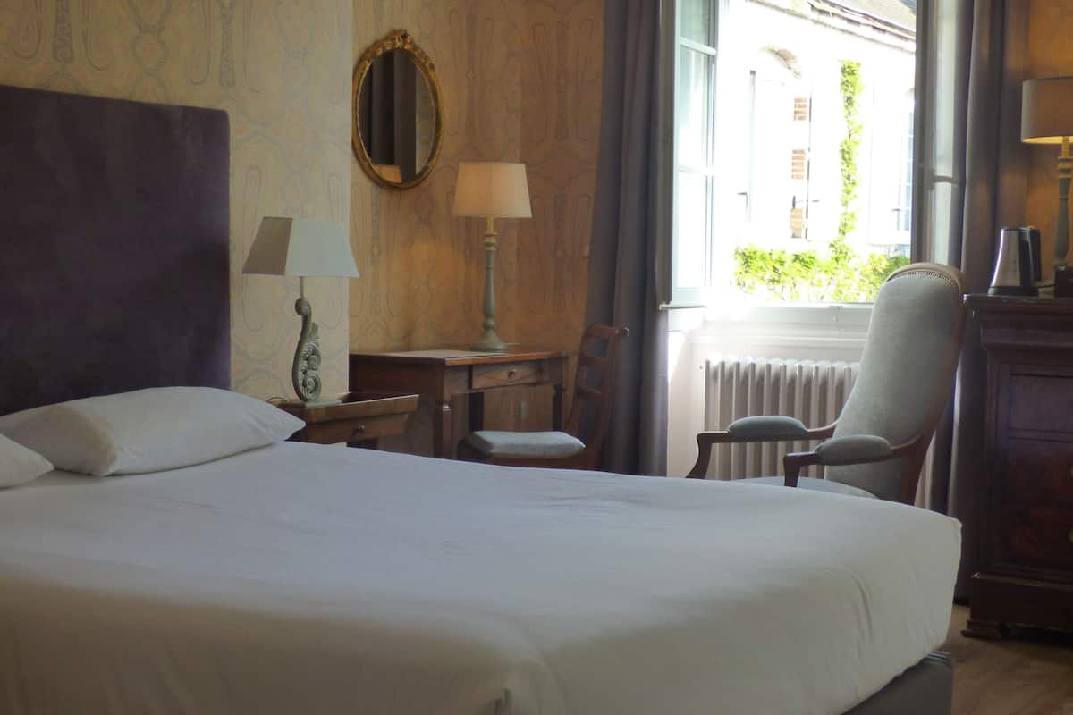 A hotel room with a large bed, a desk with a lamp, a chair, and a window with a view of buildings.
