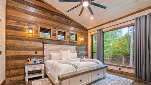 A bedroom with a wooden log wall, a large bed, a nightstand, a ceiling fan, and a window with curtains.