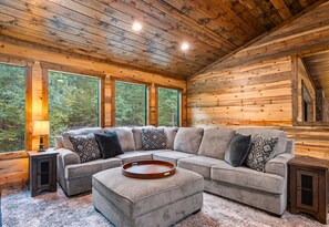 A log cabin interior with a sectional sofa, a wooden tray, and large windows offering a view of the forest.