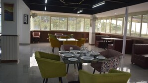 A dining area with a table set for four, surrounded by colorful chairs, large windows, and a ceiling fan. - Xenious Micro Dharmshala  (Dharamshala)