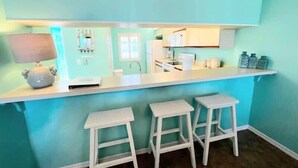 A kitchen with a teal color scheme, white countertops, and two white bar stools. - Salt Life Escape: 2B/2B in Oceanfront Complex (Cocoa Beach)