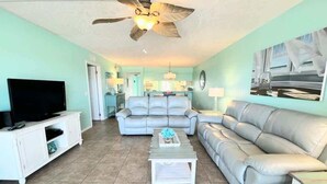 A living room with a large grey sofa, a white coffee table, a television on a white cabinet, and a ceiling fan. - Salt Life Escape: 2B/2B in Oceanfront Complex (Cocoa Beach)